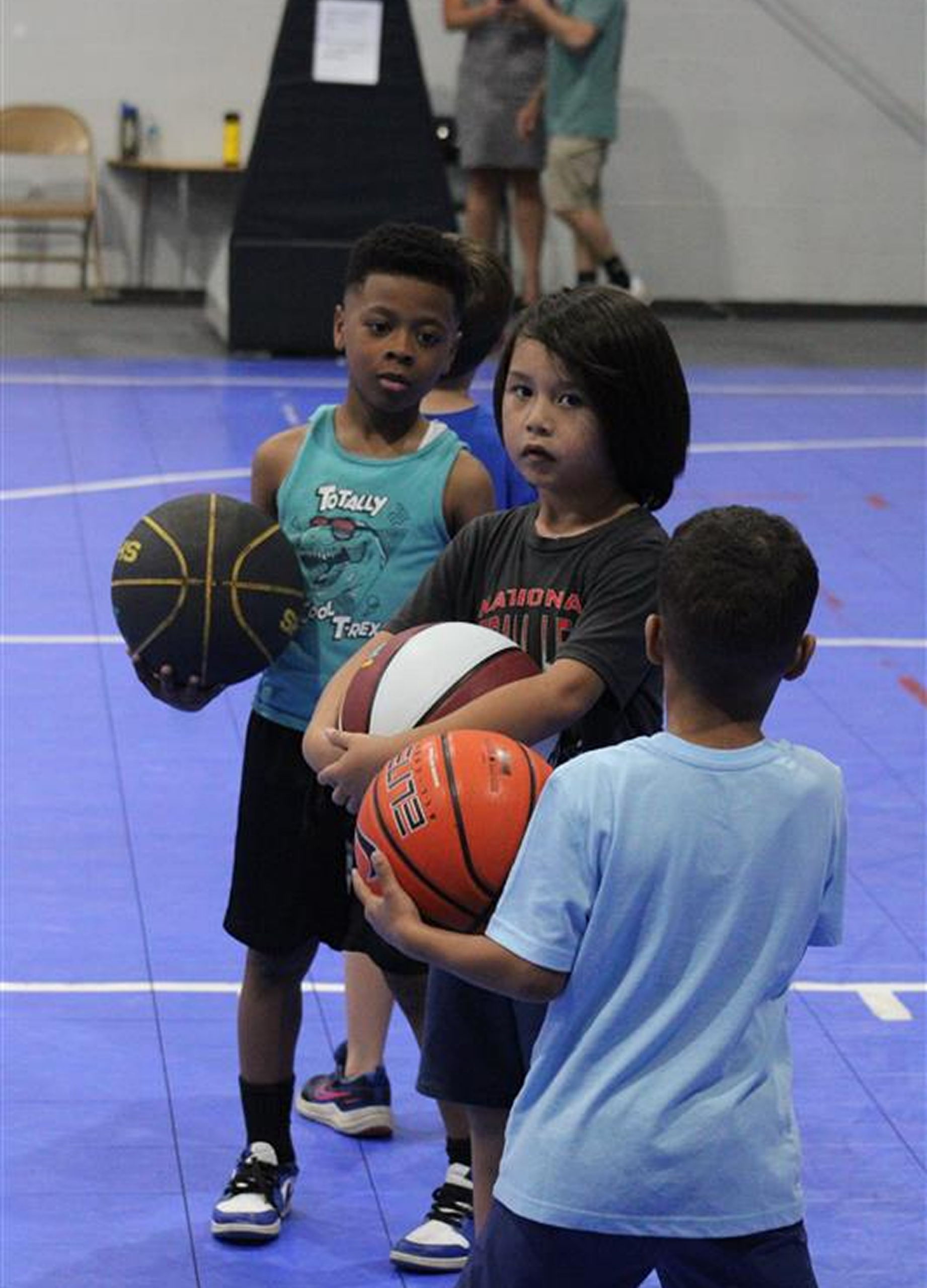 Three children stand on a blue indoor basketball court, each holding a basketball.
