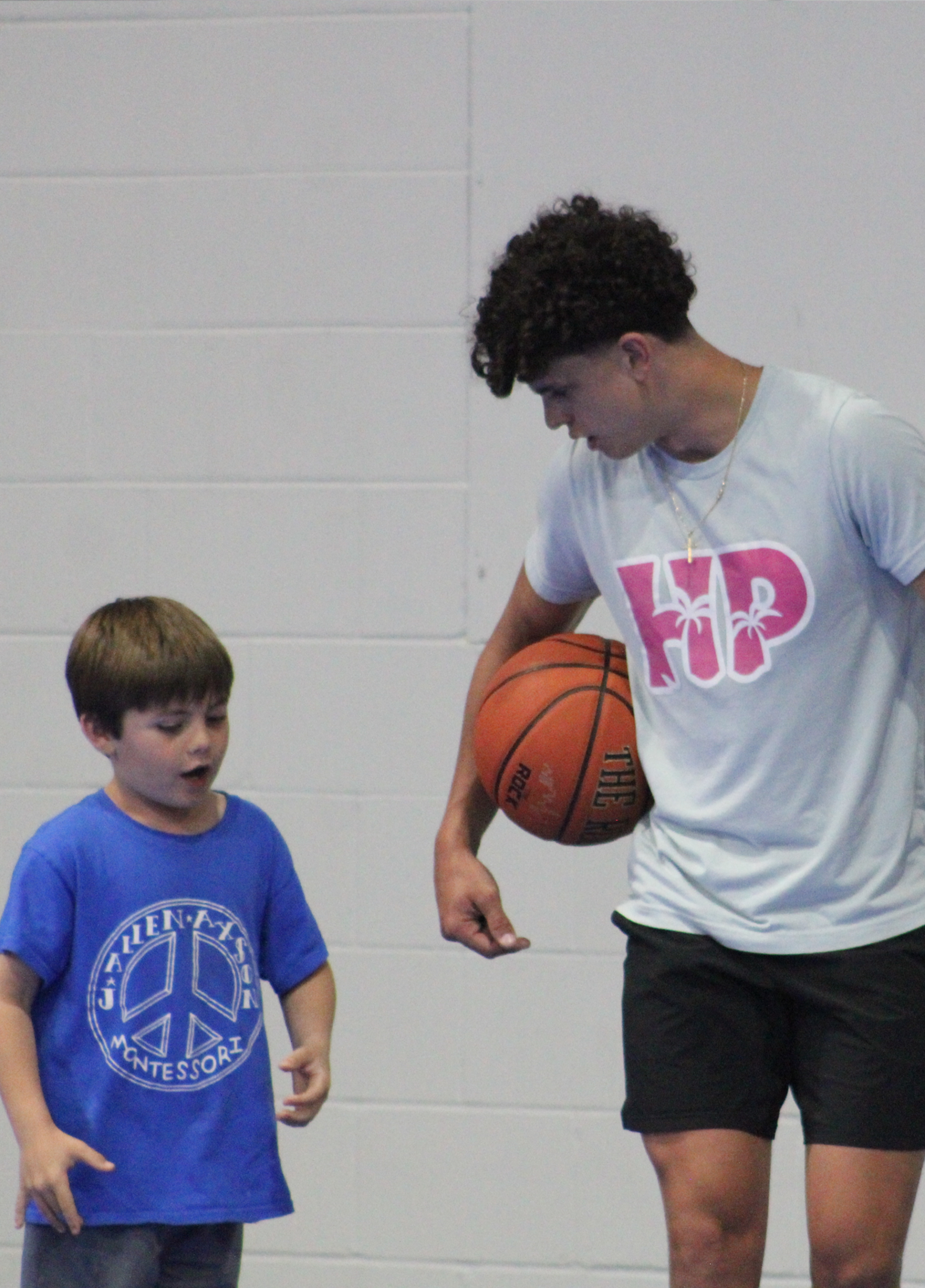 A basketball player in a light-colored shirt holds a ball while standing next to a child in a blue shirt in a gym.