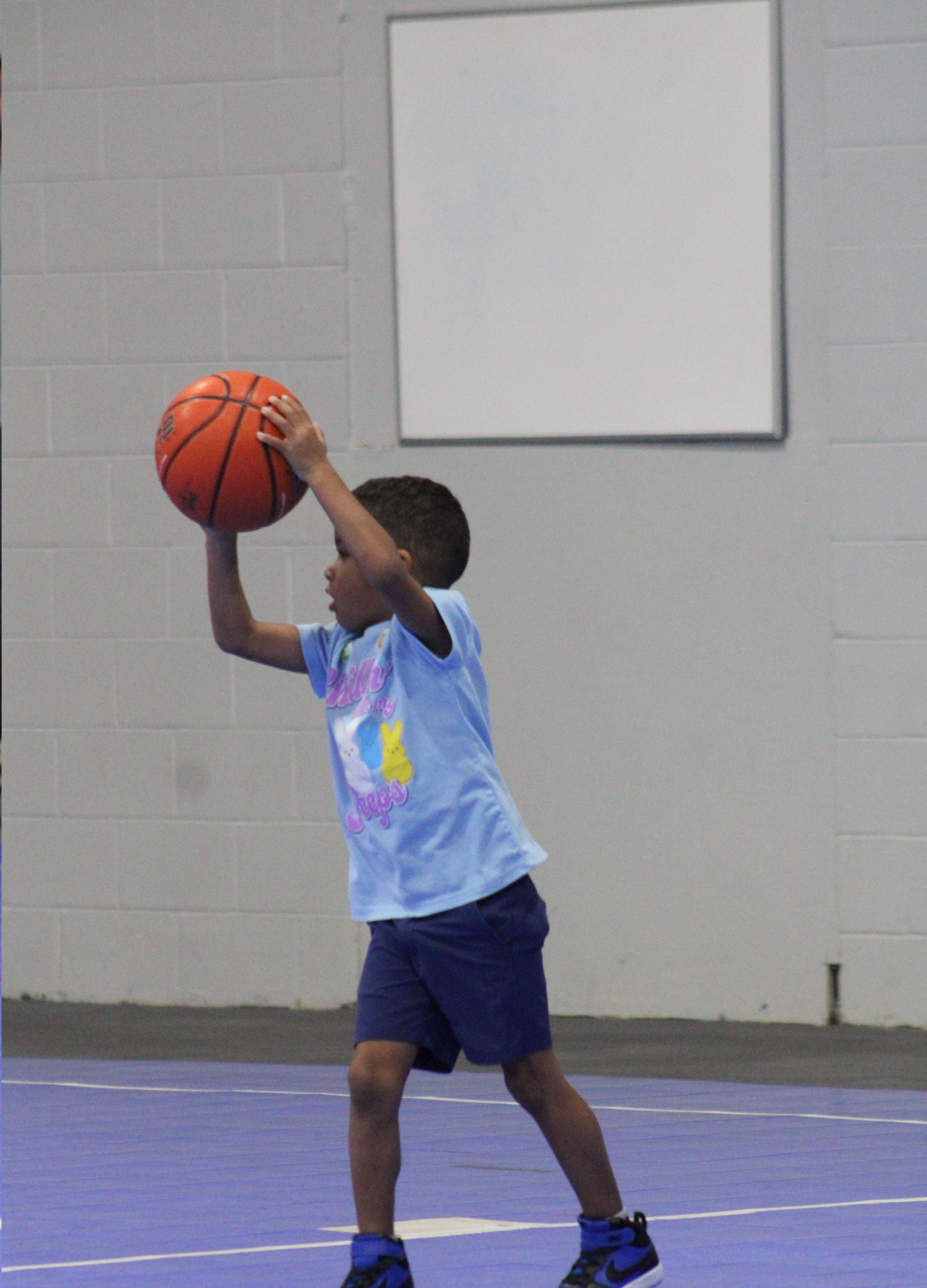 A child in a light blue t-shirt and blue shorts holding a basketball above their head on an indoor court.