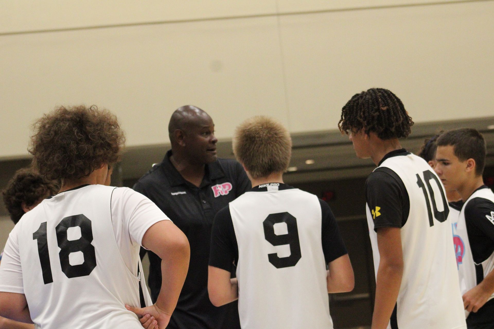A coach speaks to a group of athletes wearing white and black uniforms with numbers during a huddle in a gym.