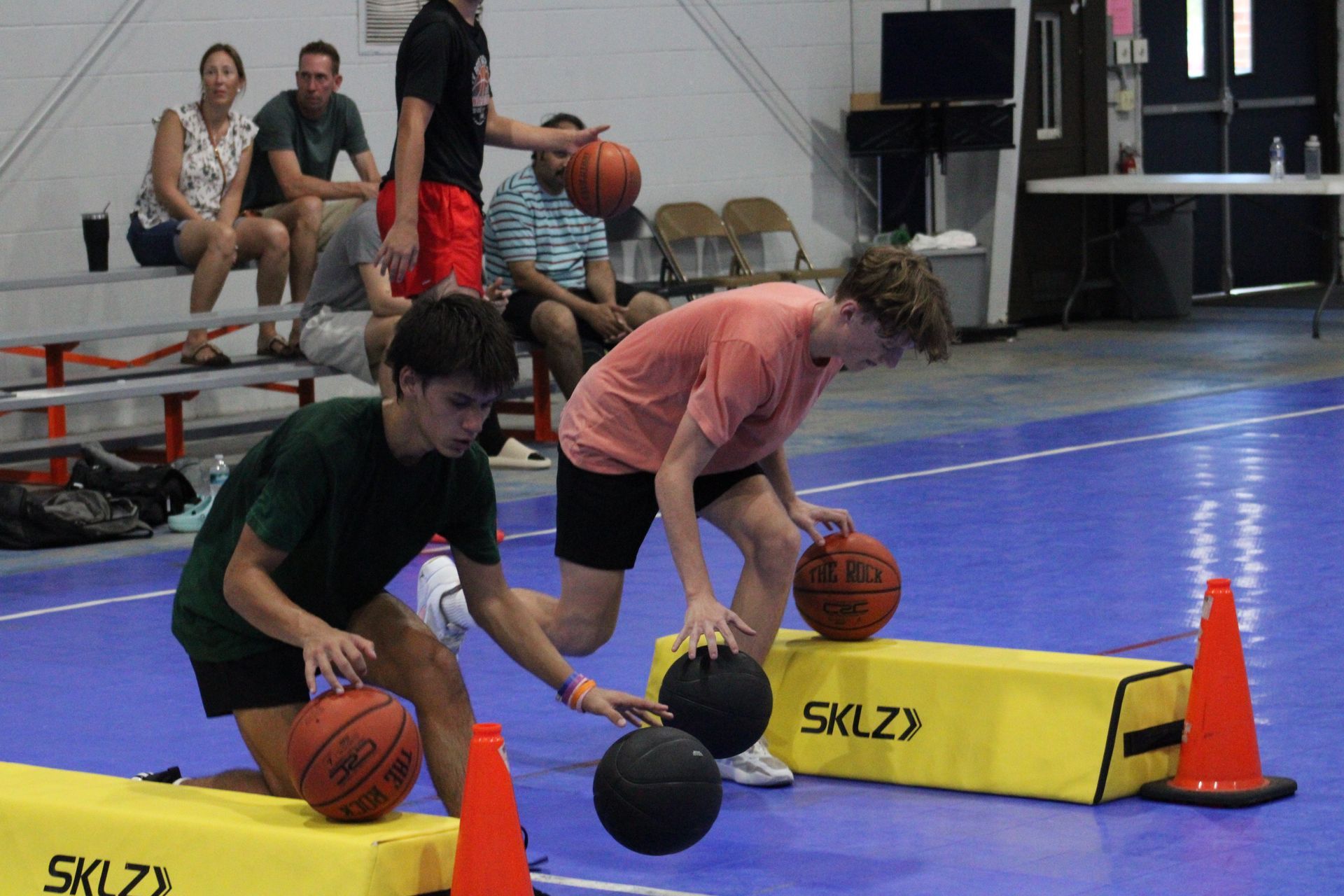 Two players dribble basketballs around yellow agility blocks and orange cones on a blue indoor court.
