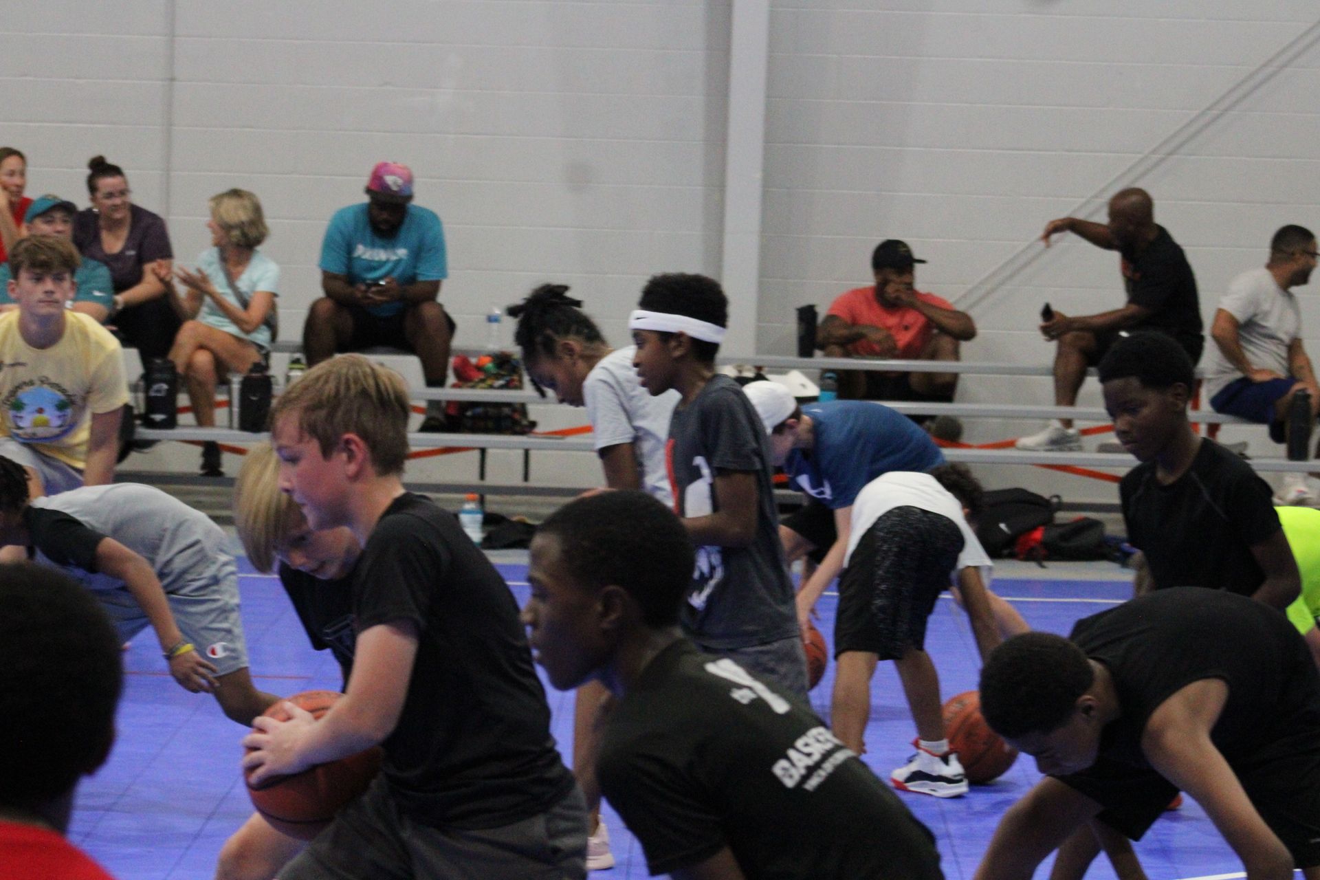 Youth athletes practicing basketball drills on a blue court with spectators seated in the background.