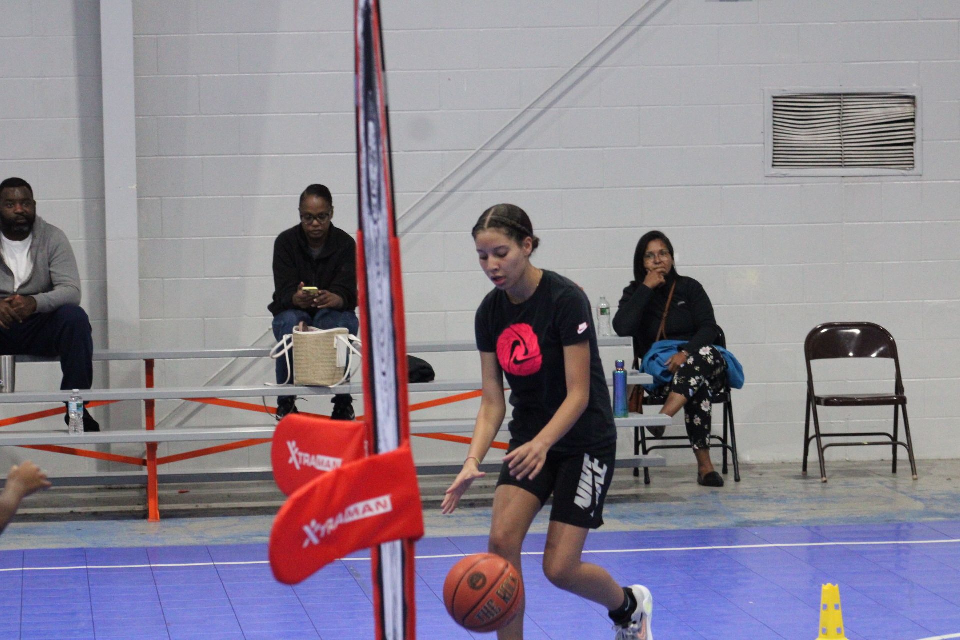 A person dribbles a basketball on an indoor blue court near a vertical pole with red pads, with onlookers in the background.