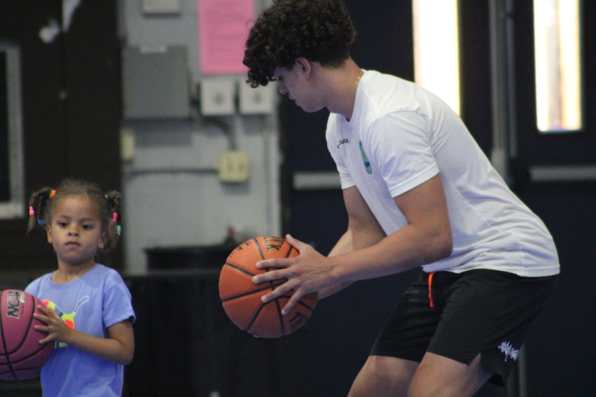 A person and a child stand side-by-side in an indoor gym, both holding basketballs while facing forward.