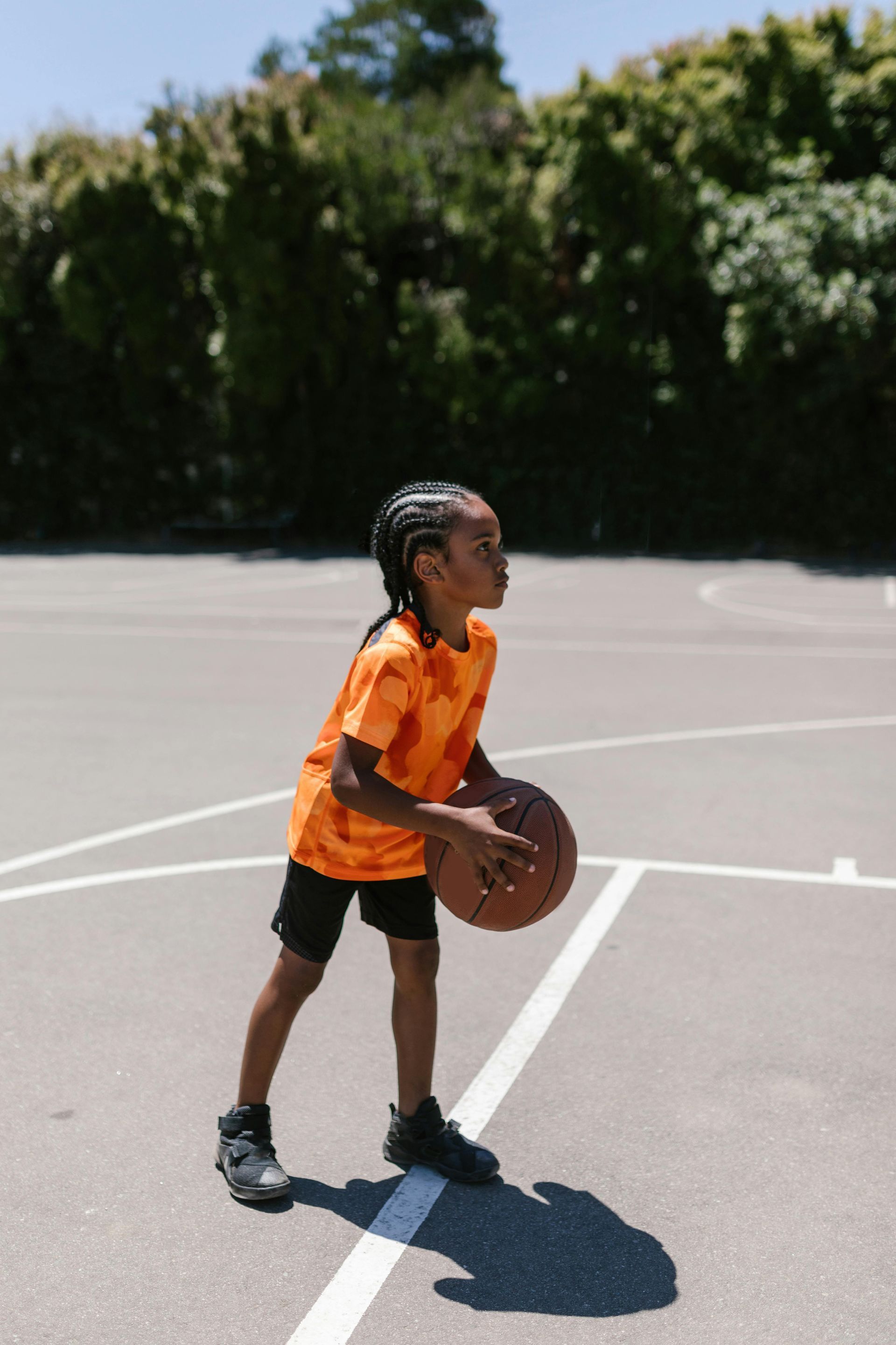 A person in an orange tie-dye shirt stands on a basketball court, holding a basketball and looking toward the right.