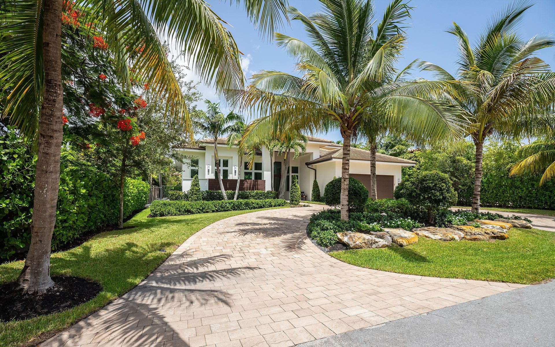 A tropical home with a brick driveway, lush landscaping, and palm trees under a sunny sky.