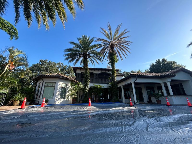 A large, light-colored house with palm trees, blue tarp in the driveway, and orange traffic cones. Bright, sunny day.