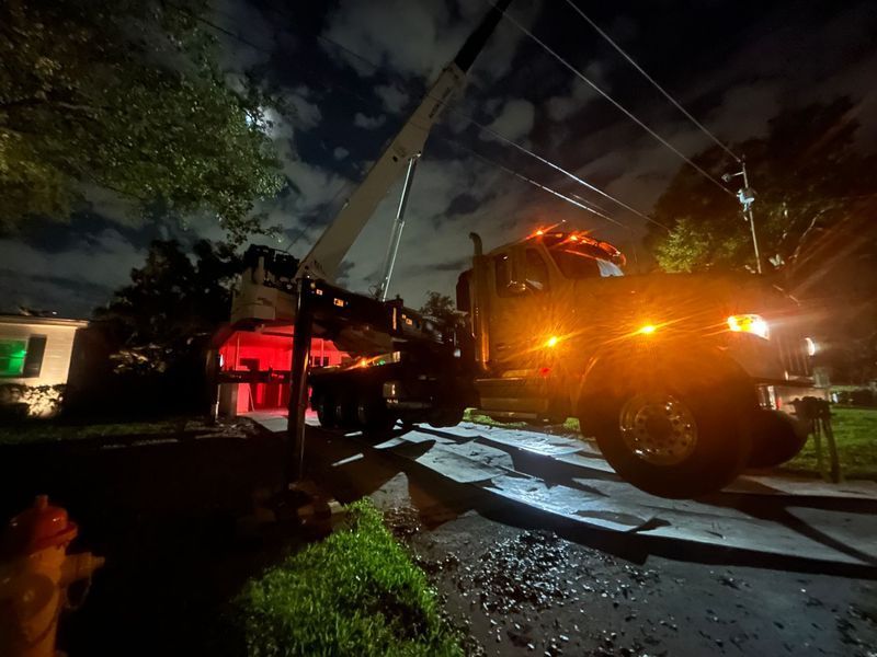 A utility truck with its boom extended works on power lines at night. The scene is lit by the truck's lights and a full moon.