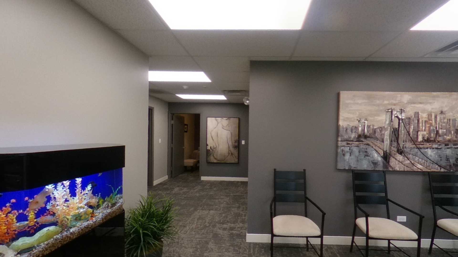 Waiting room with gray walls, artwork, chairs, and an aquarium. A hallway leads to other rooms.