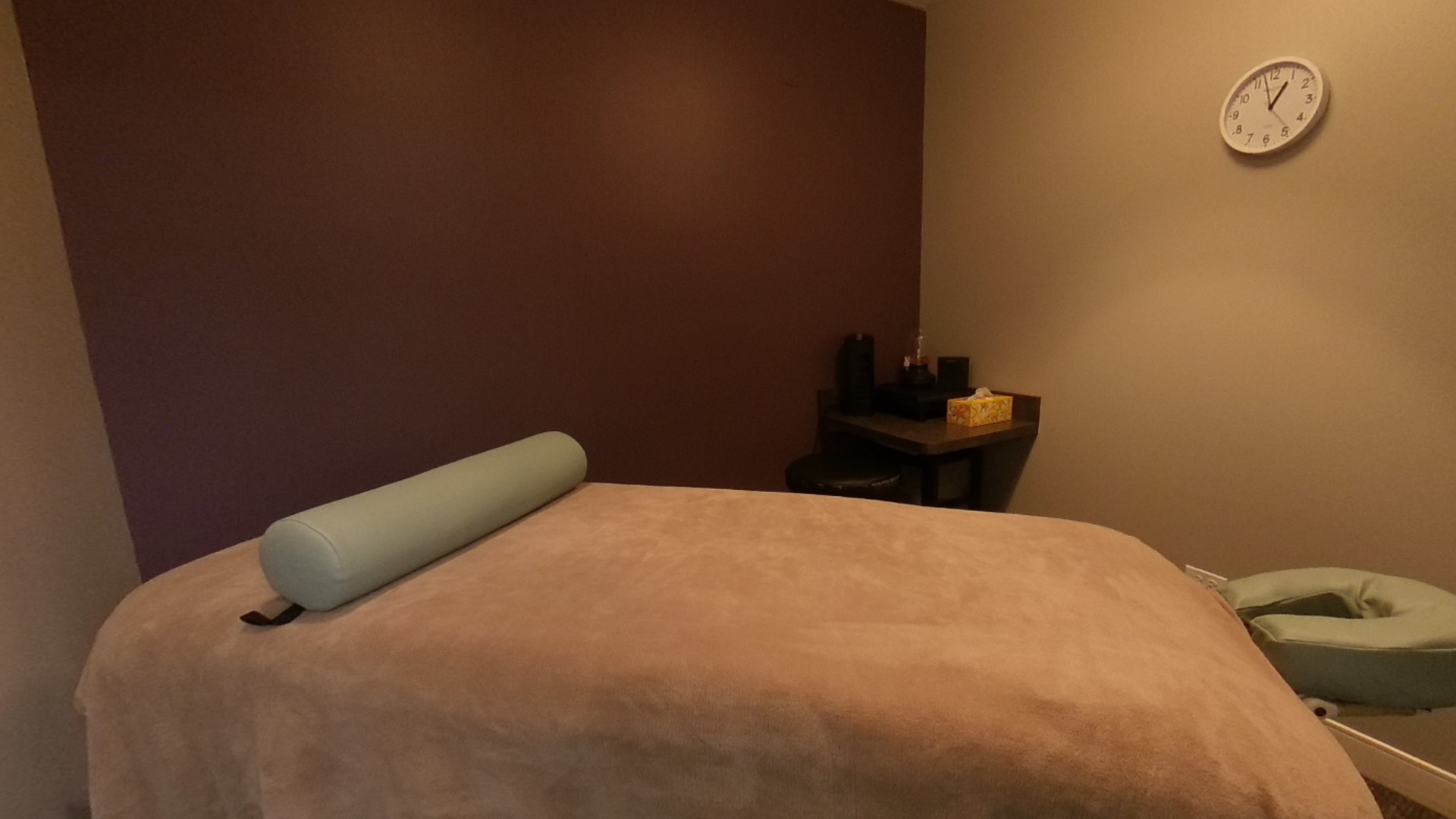 A massage room with a massage table, a cylindrical pillow, and a face cradle. There's a clock on the wall.