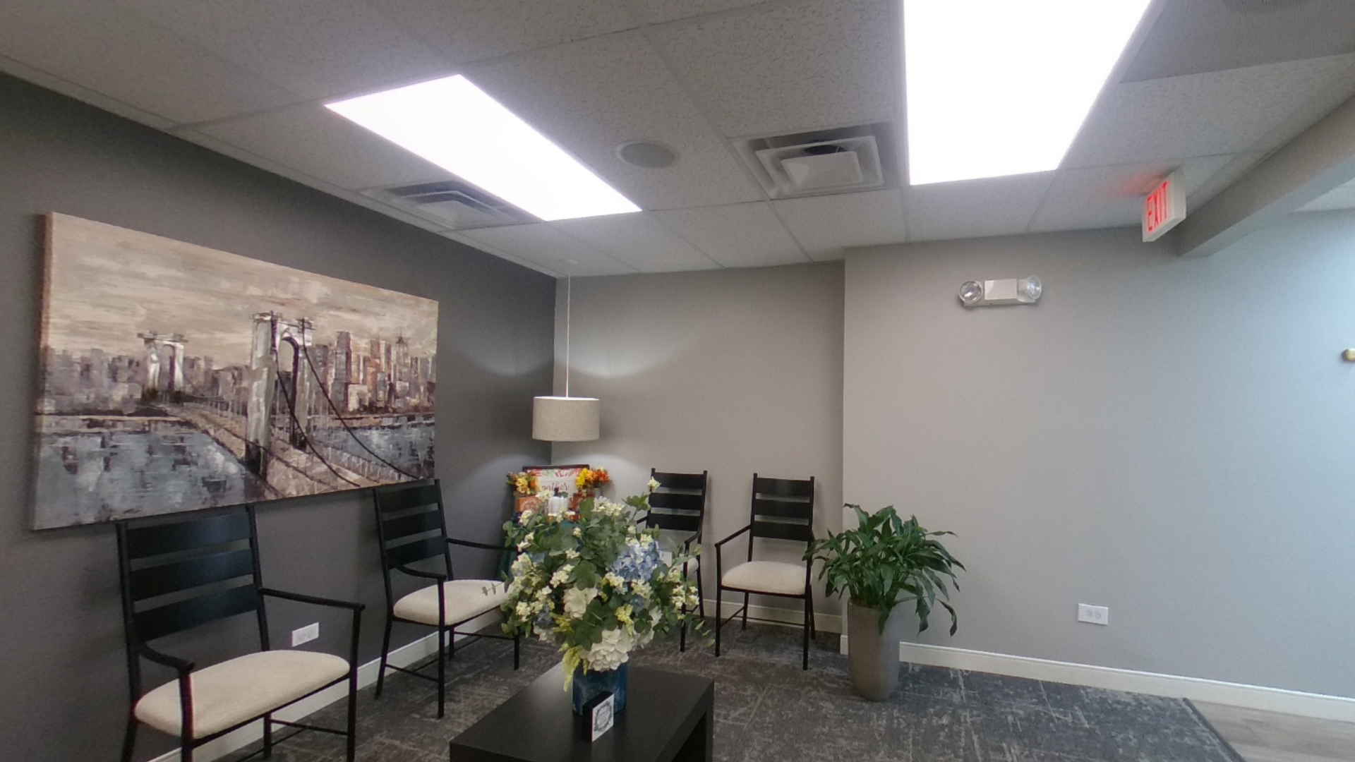 A waiting room with gray walls, black chairs, artwork, plants, and bright overhead lights.