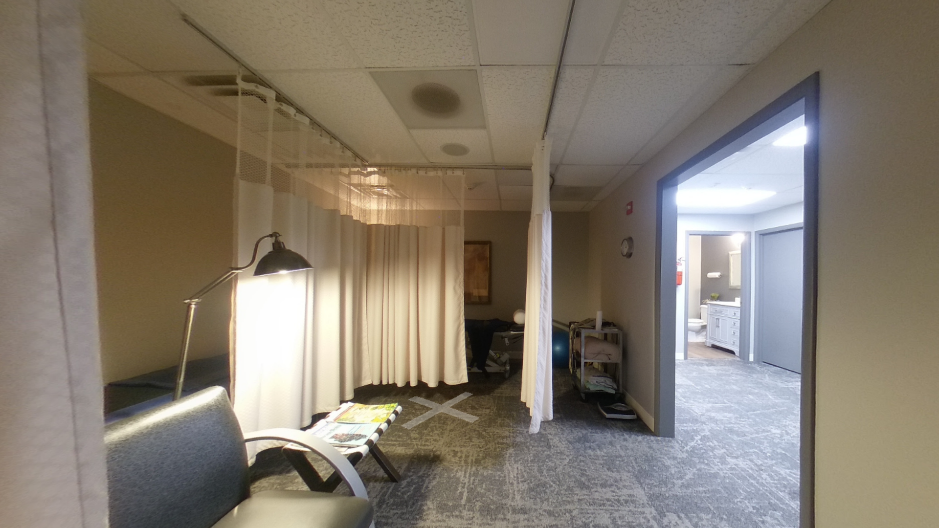 A hallway with medical examination rooms. Beige walls, gray patterned carpet, and white privacy curtains are visible.