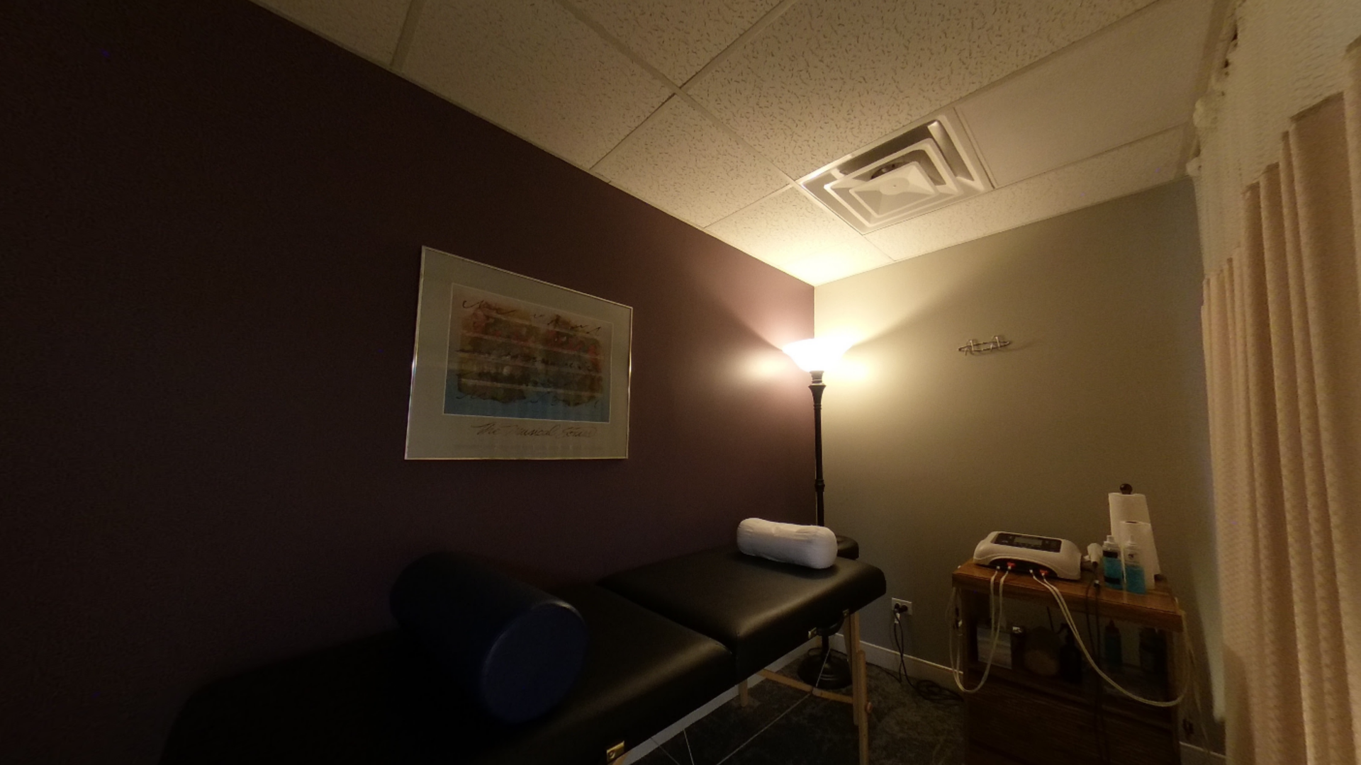Massage therapy room with a black massage table, a small wooden table with equipment, and a framed picture on a purple wall.