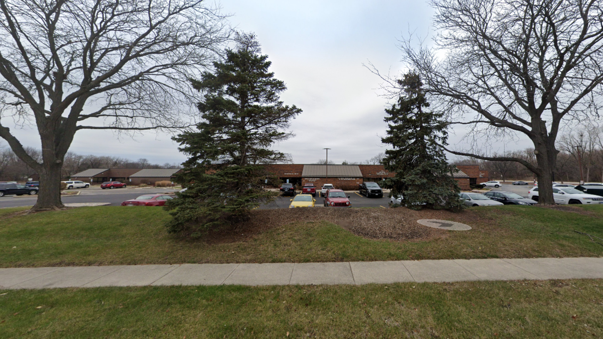 A long, one-story building with a brown facade, trees, and parked cars on a cloudy day.