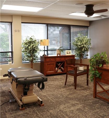 Chiropractor's office with a treatment table, a wooden cabinet, and two chairs. Large windows let in daylight; plants add greenery.