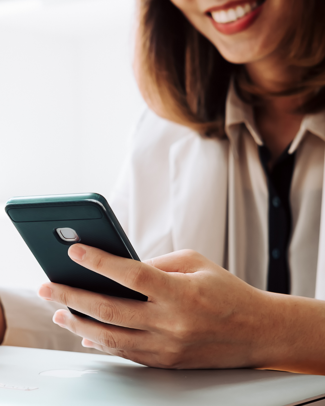 Woman smiling while looking at a smartphone in her hands. She's wearing a white blazer and sitting indoors.