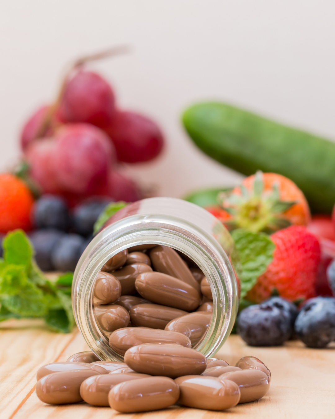 Brown capsules spilling from a glass jar, surrounded by fresh fruits like grapes, strawberries, and blueberries, and a cucumber.