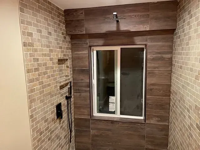 Shower with brown wood-look tiles around a window and on the ceiling. Walls have beige mosaic tile. Black shower fixtures are visible.