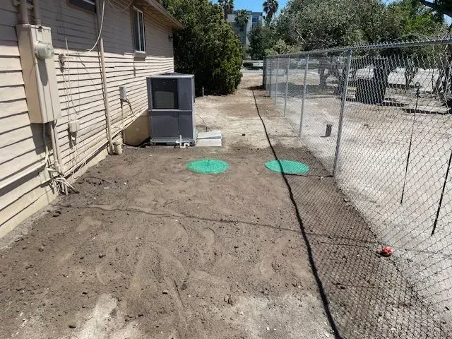 Backyard with air conditioner, chain-link fence, and two green circular markers on dirt. The ground has been recently graded.