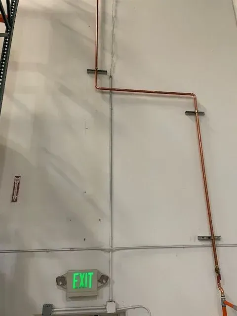 Copper pipes running along a white industrial wall, secured by metal brackets. An illuminated 