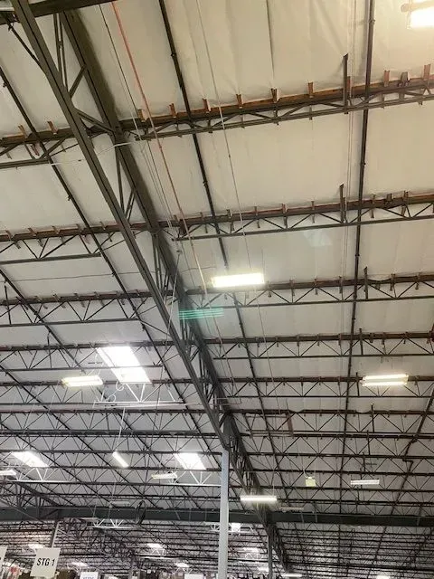 Warehouse ceiling with metal beams and insulation, featuring skylights and fluorescent lighting.