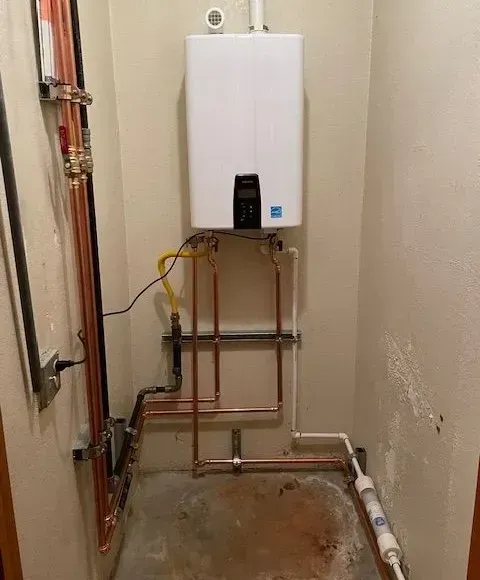 A wall-mounted, white tankless water heater with copper pipes and a water filter in a basement.