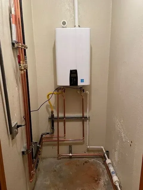 White tankless water heater mounted on a beige wall with copper plumbing pipes. It's in a utility closet with a concrete floor.