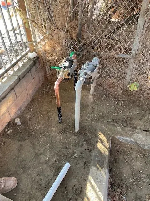 Water valves and pipes in a dirt area, near a chain link fence and low brick wall. The valves are copper and white, with green handles.