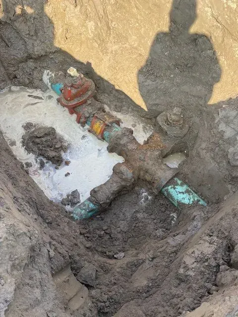 A rusty, old water pipe in a trench, with a red valve visible. A person's shadow is cast on the dirt.