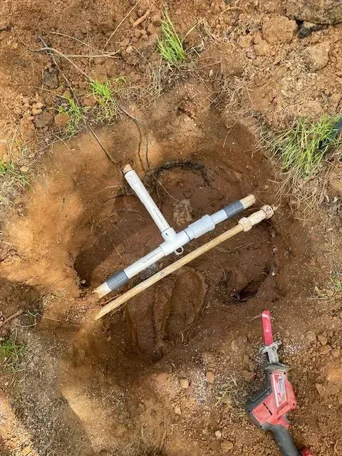 A hole in the dirt reveals a plumbing apparatus with PVC piping and a drill. Outdoor setting.