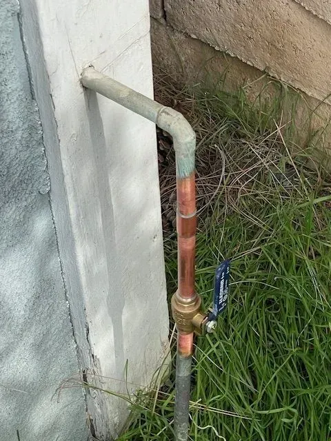 Copper water pipe with a blue-handled valve, attached to a wall and ground. Green grass surrounds the pipe.