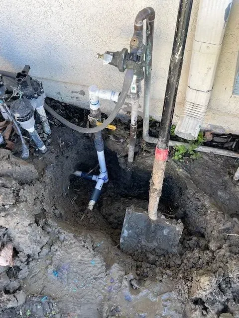 Hole dug in dirt next to a house, revealing irrigation system valves and pipes. A shovel leans against the edge of the hole.