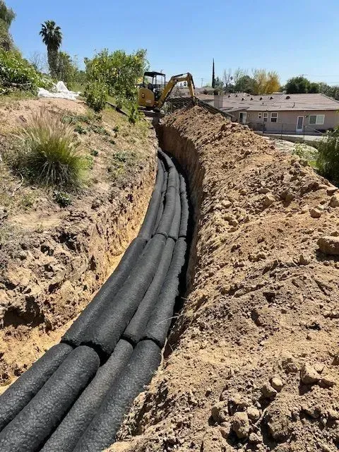 A long trench filled with black, cylindrical structures. A yellow excavator works nearby on a sunny day.