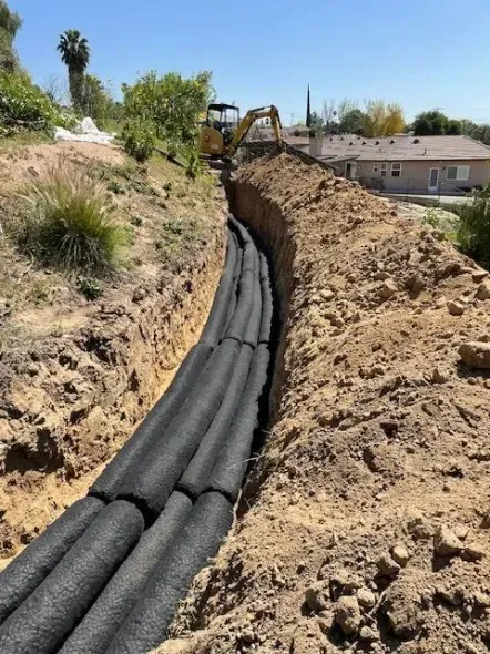 A trench with black tubes is being dug. A backhoe is visible in the background, with residential houses and vegetation nearby.