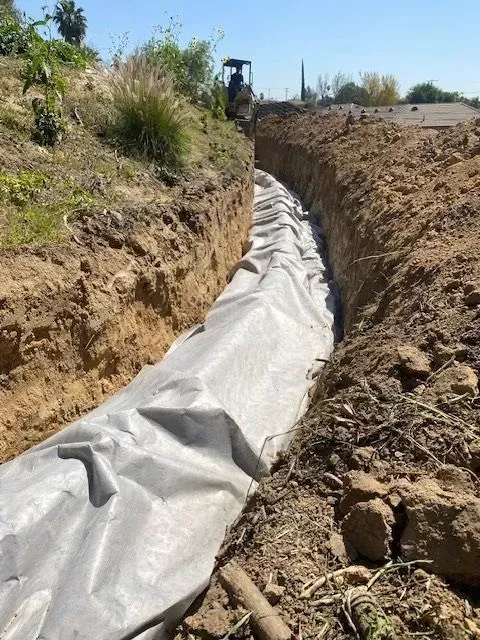 A long trench lined with gray fabric in an earthen ditch; an excavator is in the background.