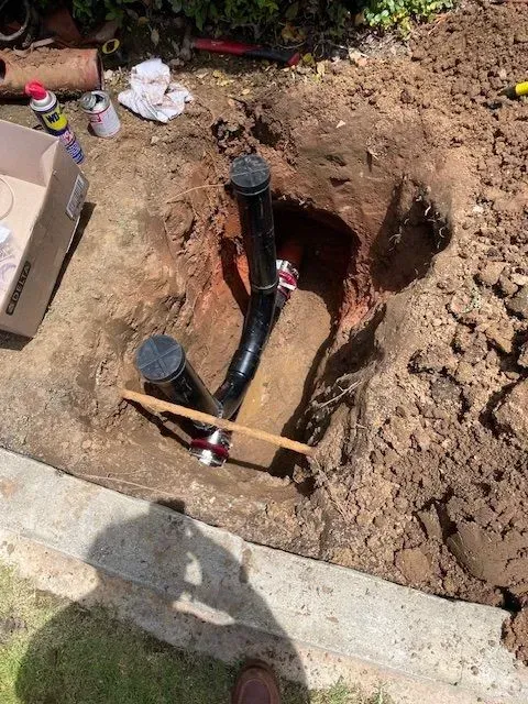 Excavated dirt reveals black pipes, likely plumbing, in a yard. Tools and materials surround the hole, suggesting repair or installation.