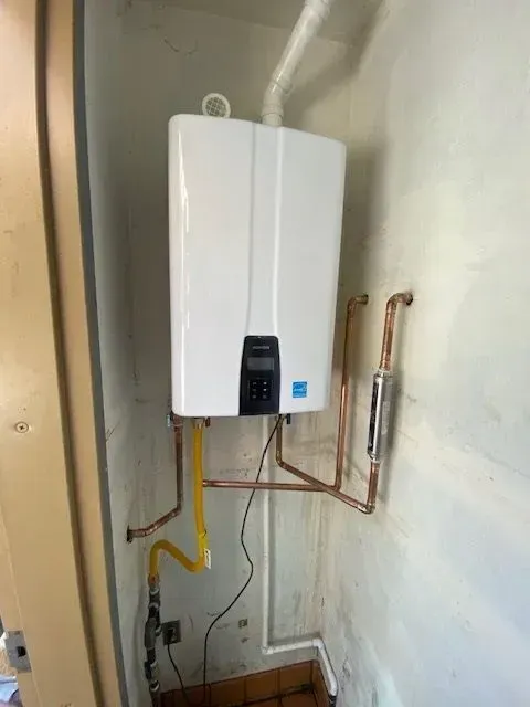 A white tankless water heater installed in a corner, with copper pipes and a yellow gas line.