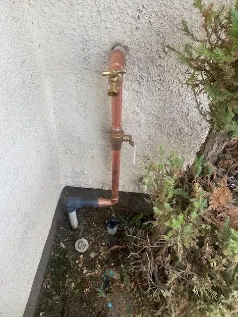 Copper pipes with two spigots attached to a stucco wall near a bush and a water valve cover.