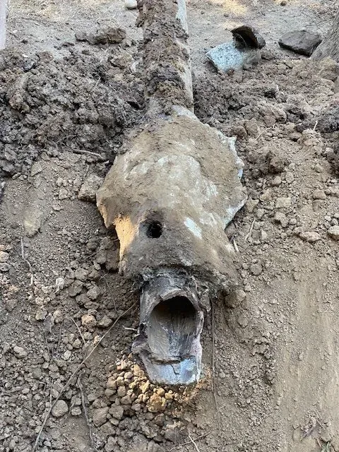 Severed section of pipe in dirt, partially covered in concrete, with a hole at the top and open bottom end.