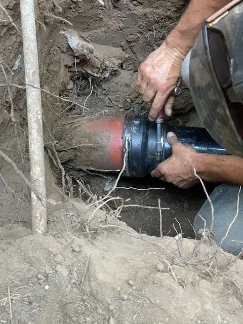 A person is connecting a black pipe with a red fitting in a dirt trench. The person uses a wrench on a clamp.