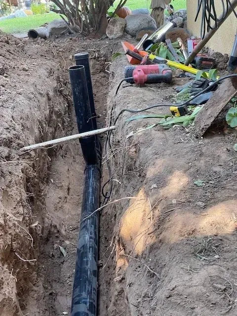 A trench holds black pipes for an underground installation. Tools are scattered on the edge, and the surrounding soil is brown.