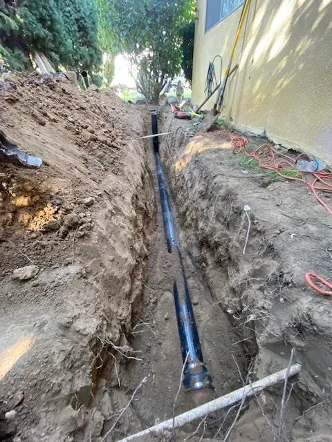 A trench dug near a yellow building, revealing black pipes laid in the earth. The dirt sides are visible, with tools and wires nearby.