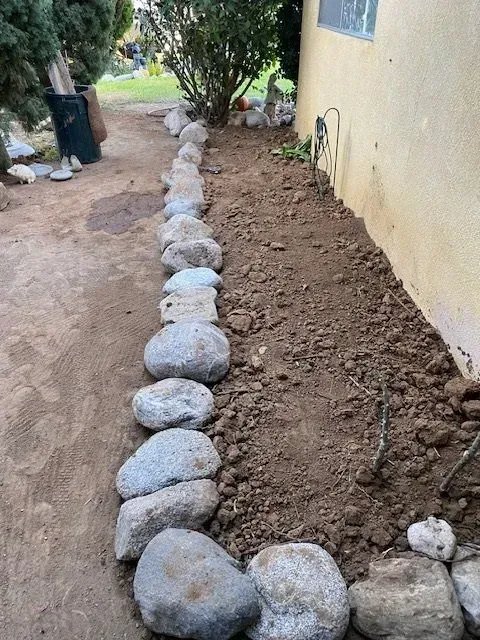 A rock border lines a dirt garden bed next to a stucco wall. Brown dirt and concrete ground surround the border.
