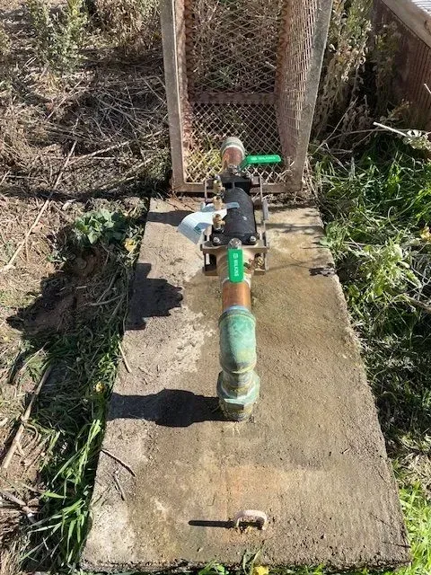 Green-handled water spigot with pipes and gauge on a concrete pad, near a chain-link fence, outdoors.
