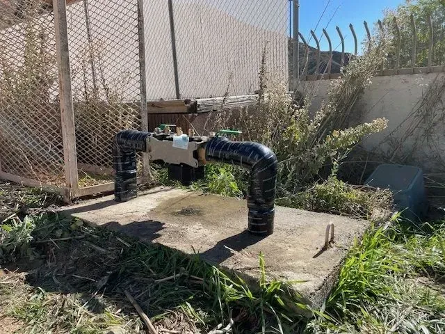 Water pipe with valve on a concrete base, wrapped in black tape. Set in a grassy area near a fence.