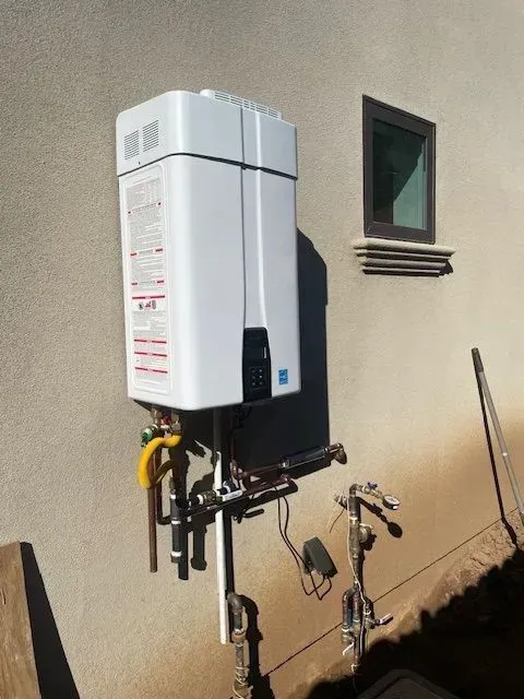An outdoor, white tankless water heater mounted on a light-colored stucco wall next to a small window. Copper piping and gas lines are visible below.