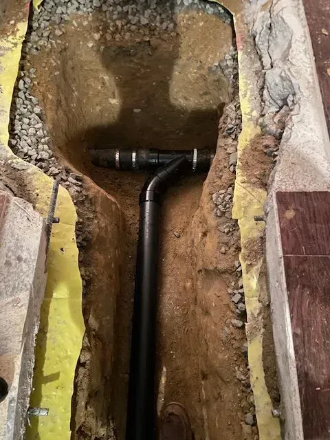 Trench dug in a floor, revealing black plumbing pipes with fittings and gravel, surrounded by yellow tape and concrete.