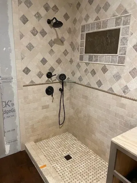 Shower stall with beige tile, diamond pattern on top, and a tiled ledge. Black shower fixtures installed.