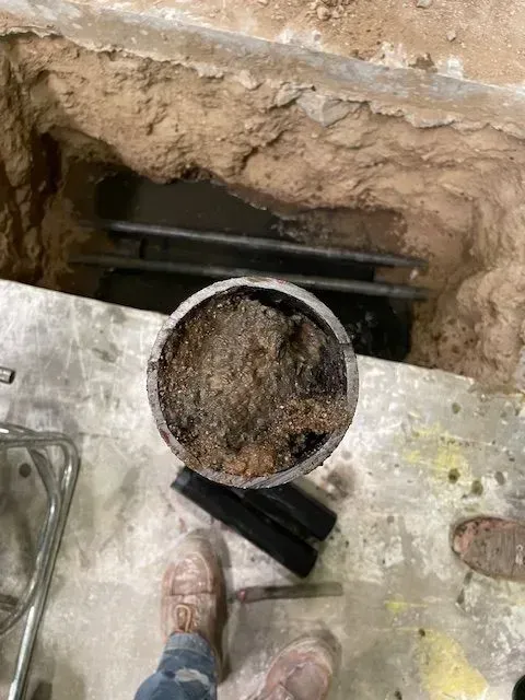 A hole with a large pipe filled with debris. Two black pipes and a pair of feet are also visible.