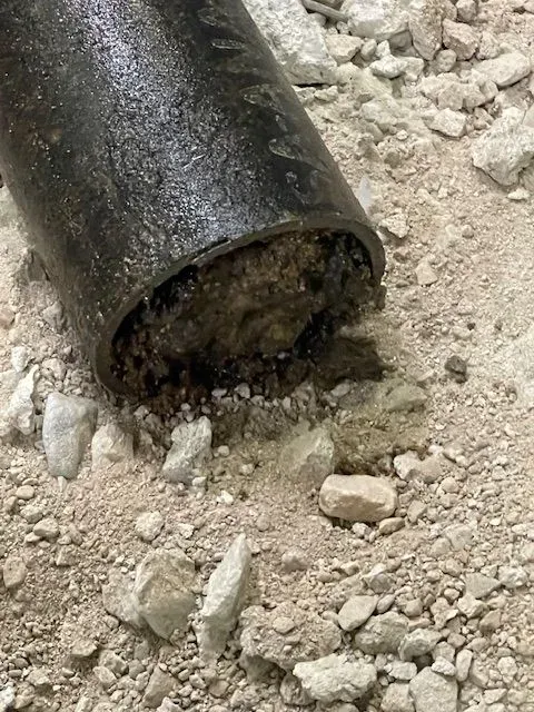 Cut open, rusty metal pipe filled with hardened sediment on a bed of gravel and sand.
